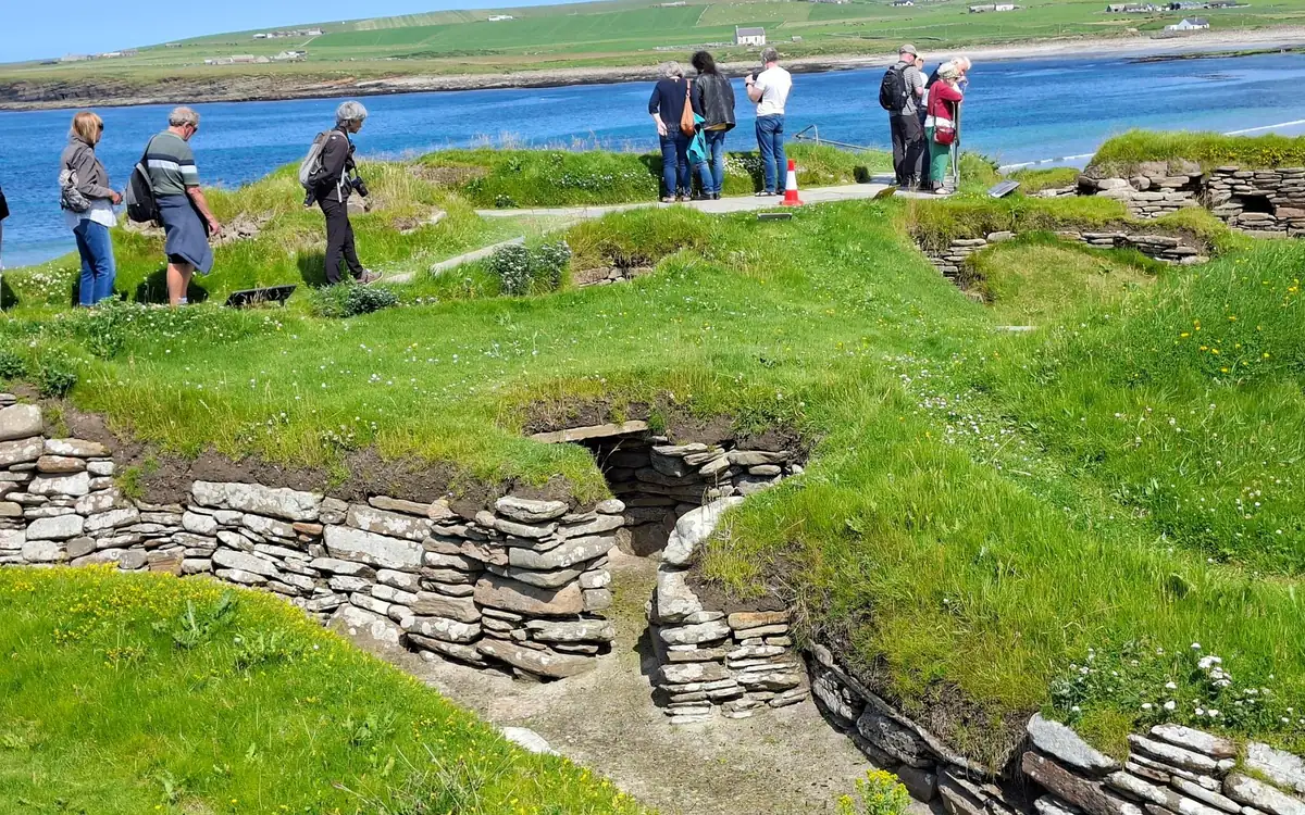 Visitors on the path above the sunken stone houses of a Neolithic village by a sandy bay