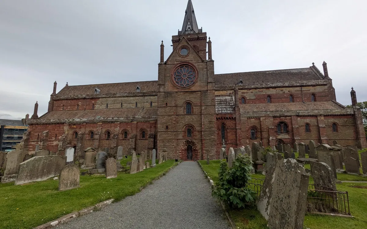 A red sandstone cathedral with a rose window and a spire behind a graveyard