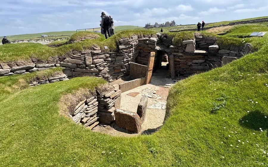 Stone-walled houses sunk into green turf, visitors on the path above and a large house behind