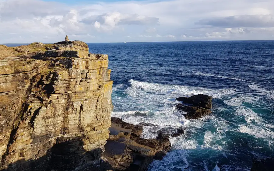 A layered sandstone sea cliff above white surf, a small stack of stones on top