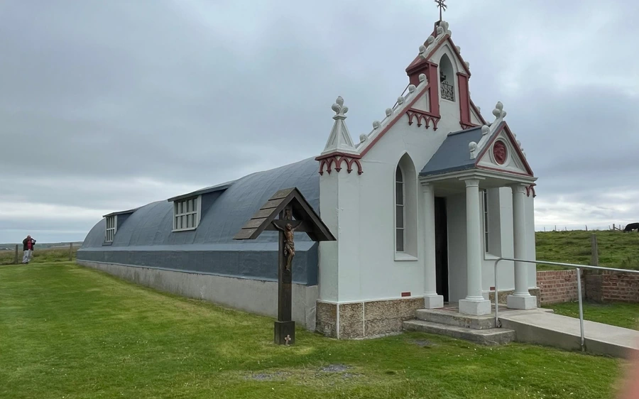 A small white chapel with red trim built onto a curved grey hut, on a green lawn